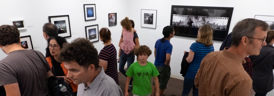 A crowd of art lovers enjoying a past Somerville Toy Camera Fest exhibition at the Brickbottom Gallery. A variety of framed black & white and color photos are visible lining the walls.