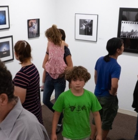 A crowd of art lovers enjoying a past Somerville Toy Camera Fest exhibition at the Brickbottom Gallery. A variety of framed black & white and color photos are visible lining the walls.
